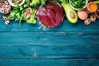 Overhead view of a blue surface with various foods high in vitamin B9, including liver, avocado, broccoli, spinach, parsley, beans, and nuts.