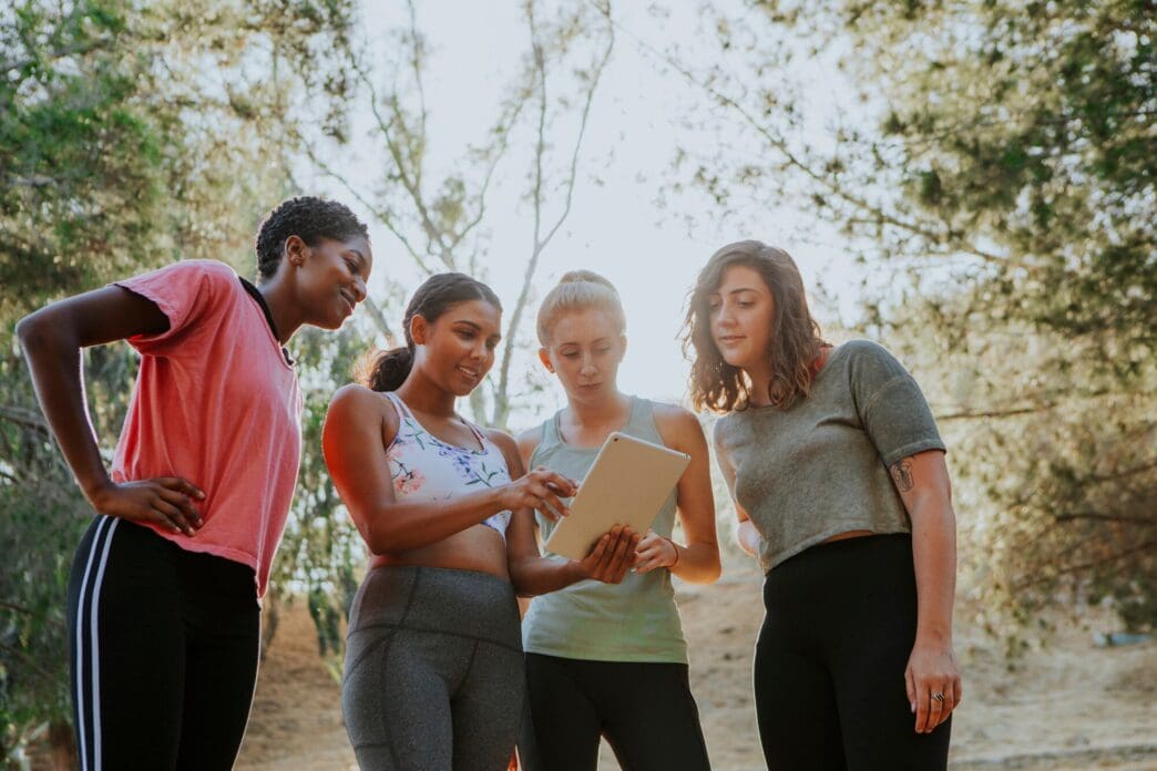 A group of women, engaged in an outdoor activity, are looking at a digital tablet.