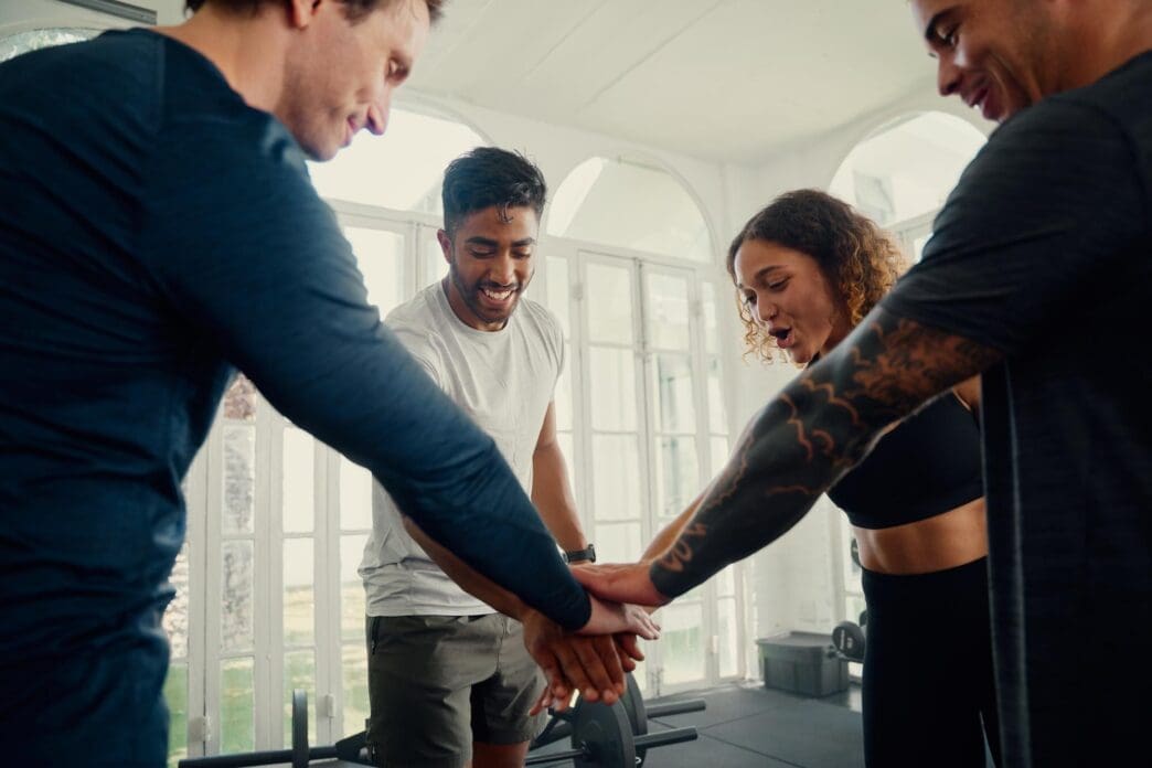 A group of young adults of diverse ethnicities in athletic wear smile and hold their hands together in a gym.