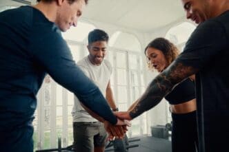 A group of young adults of diverse ethnicities in athletic wear smile and hold their hands together in a gym.