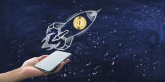 A hand holds a smartphone displaying a Bitcoin symbol launching upward against a chalkboard background, illustrating cryptocurrency investment.