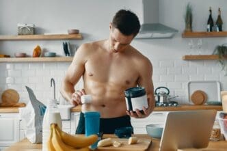 A muscular man stands in a kitchen, preparing a protein drink.
