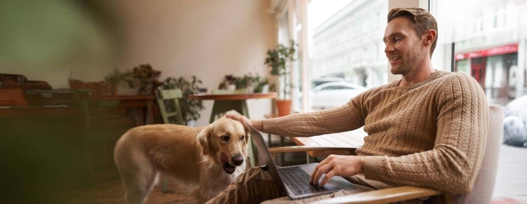 A young man pets his golden retriever while working on a laptop in a dog-friendly cafe.