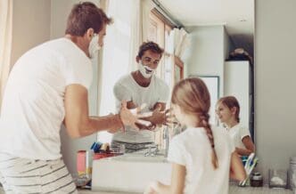 A young child and their father are in a bathroom, smiling as they share a moment while shaving in front of a mirror.