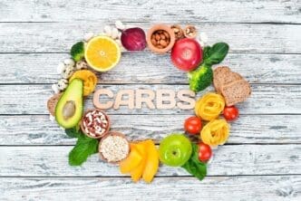 Overhead shot of a white surface showcasing various healthy carbohydrate-rich foods, including bread, pasta, avocados, flour, pumpkin, broccoli, beans, and spinach, with ample copy space.