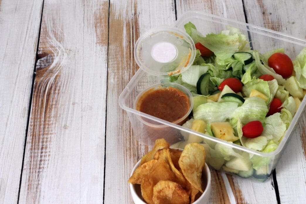 A transparent box contains a healthy meal of fresh vegetable salad and crispy chips.