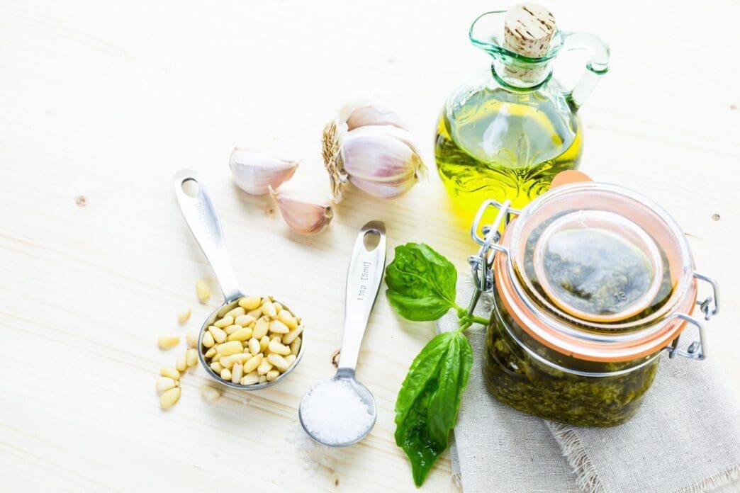 A close-up shows a bowl of vibrant green homemade basil pesto sauce surrounded by fresh basil leaves, pine nuts, and garlic.