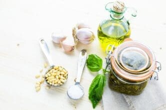 A close-up shows a bowl of vibrant green homemade basil pesto sauce surrounded by fresh basil leaves, pine nuts, and garlic.