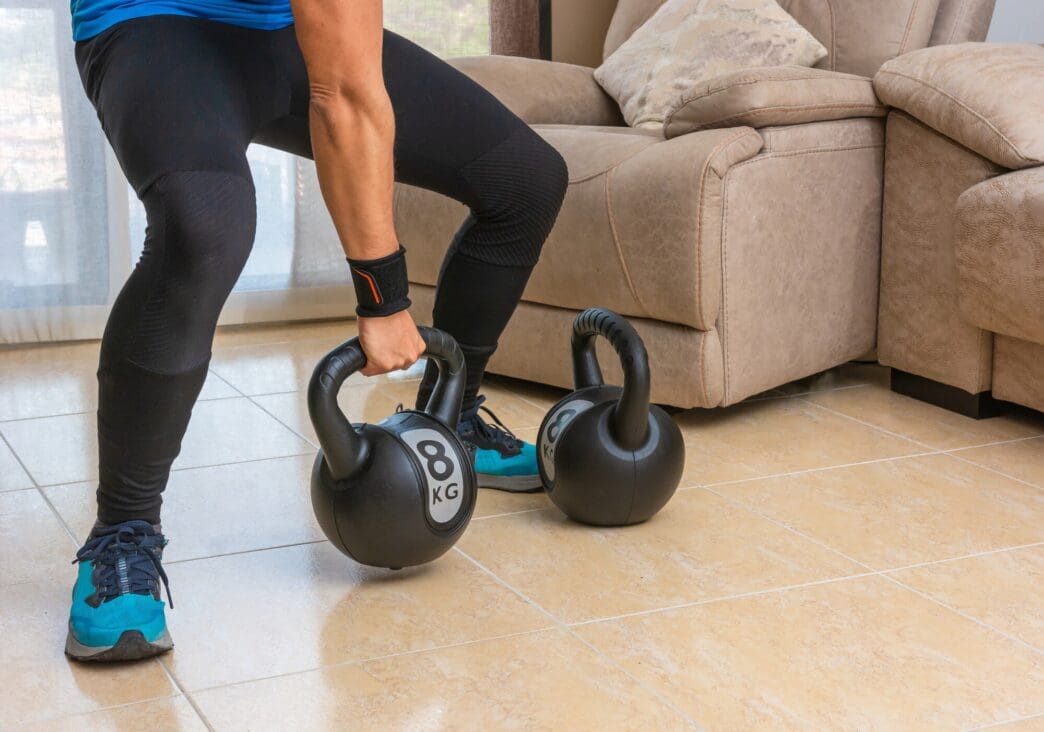 A Latin man in athletic wear exercises at home using two kettlebells.