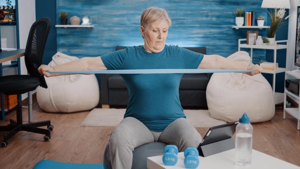 An elderly woman exercises on a toning ball, pulling a resistance band while watching a workout video on a tablet.