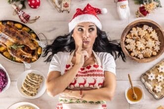 A woman in a Santa hat and Christmas apron lies on the floor surrounded by gingerbread cookies, gazing to the side, with a roast goose and side dishes nearby.