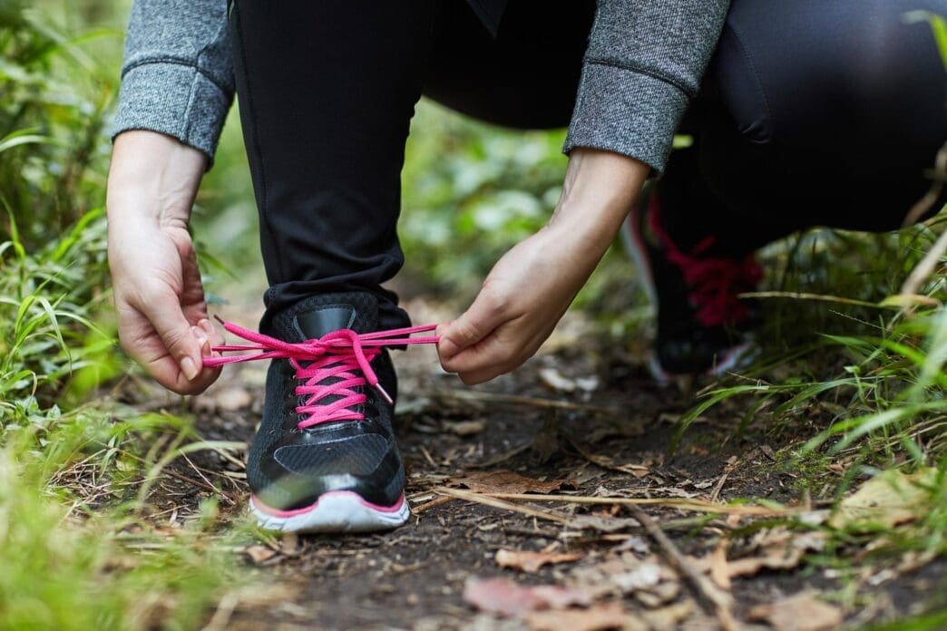 Person tying a shoelace on a sneaker while hiking on a forest trail.