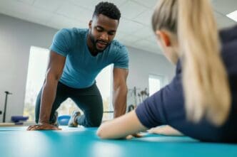 A personal trainer assists a young Caucasian woman in a fitness class, both positioned on a bright blue mat.