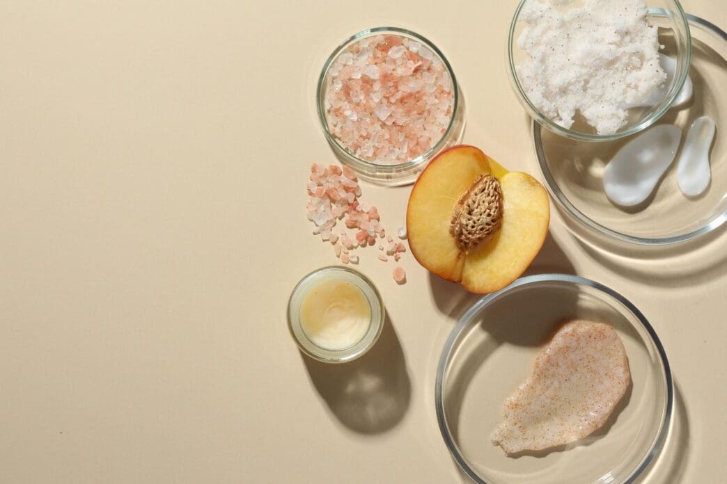 Flat lay of petri dishes filled with various cosmetic products and a peach, arranged on a beige background with space for text.