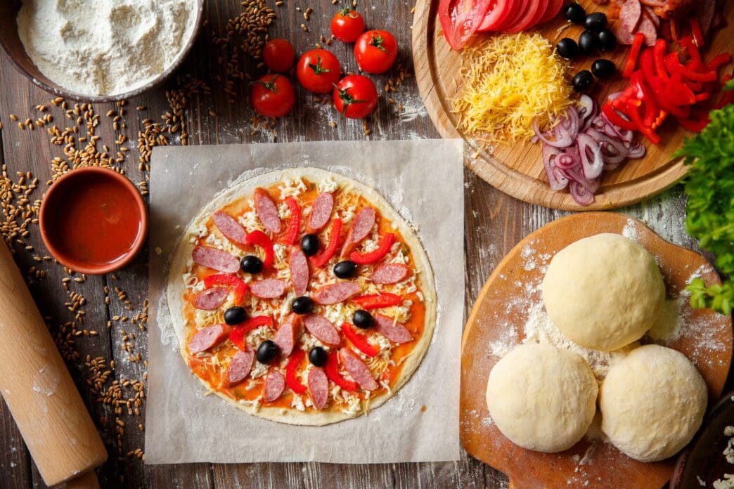 Pizza dough on a wooden surface is topped with various pizza ingredients.
