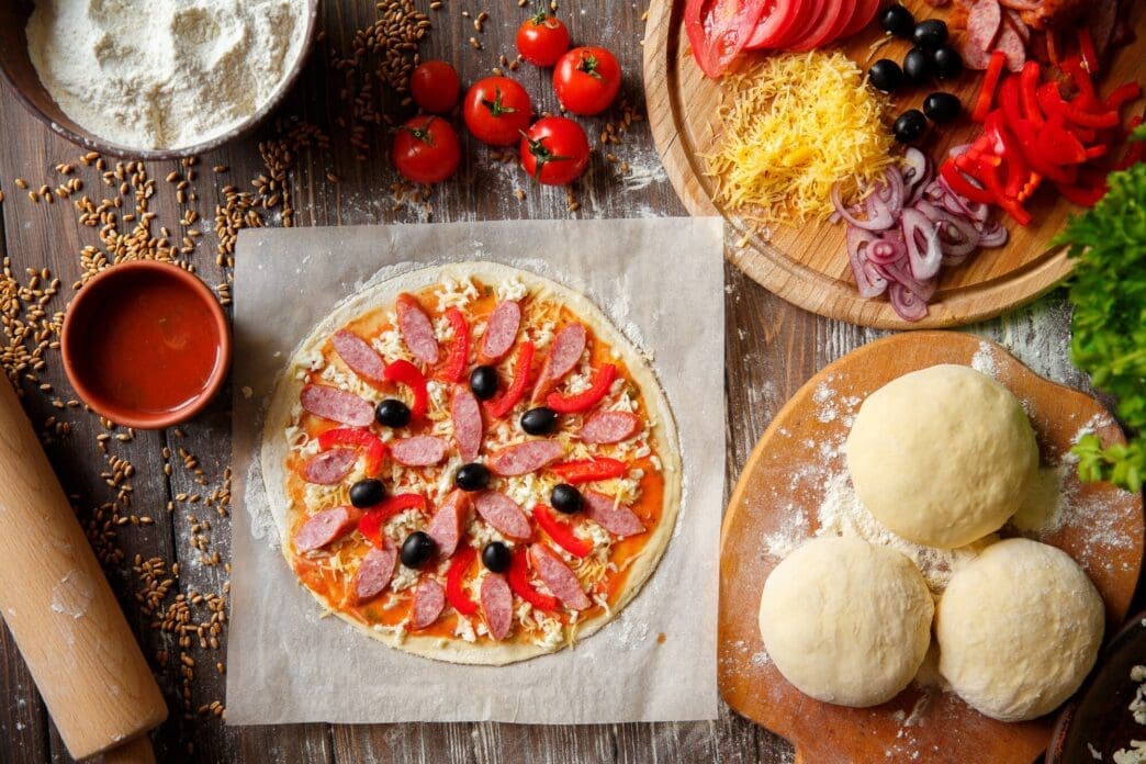 Pizza dough on a wooden surface is topped with various pizza ingredients.