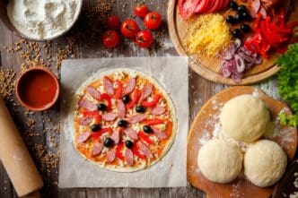 Pizza dough on a wooden surface is topped with various pizza ingredients.