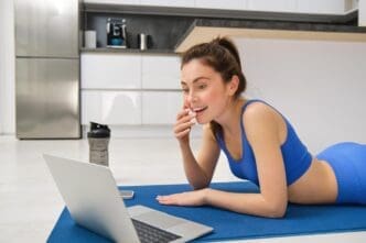 A smiling woman sits with a laptop, appearing to watch a fitness activity.