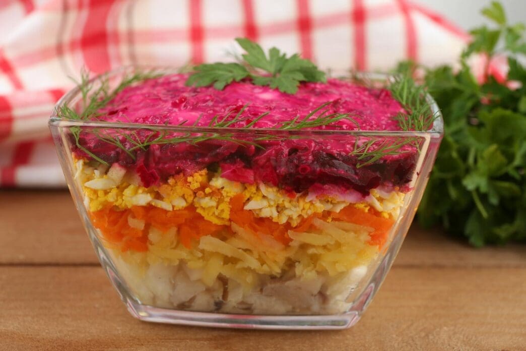 A glass bowl of Russian herring salad sits on a wooden table.