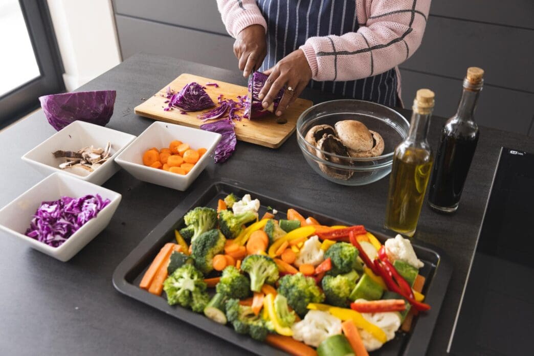 Senior biracial woman in an apron cuts cabbage on a kitchen table surrounded by vegetables.