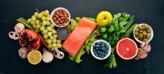 Overhead view of a spread of healthy foods, including fish, nuts, protein, berries, vegetables, and fruits, arranged on a black wooden surface with space for text.
