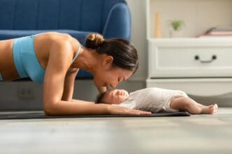 An Asian mother in workout clothes does a plank exercise and kisses her baby on a yoga mat.