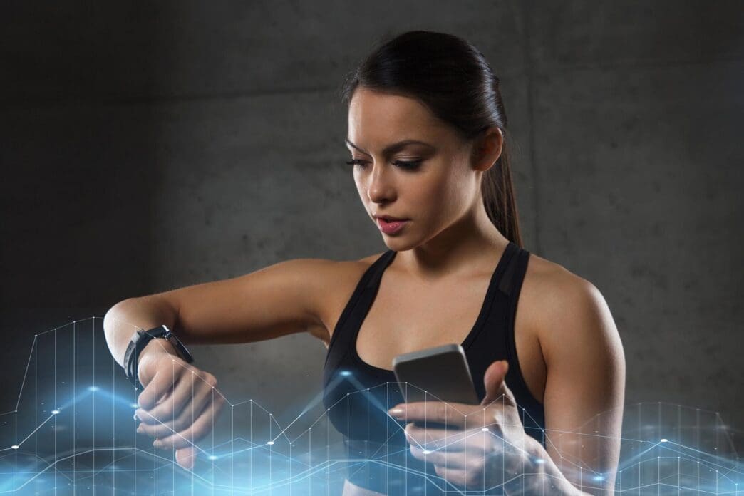 A young woman in a gym uses a smartwatch and smartphone to track her workout.
