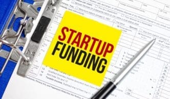 A yellow sticky note displays a text message about startup funding, resting atop a file folder containing tax forms.