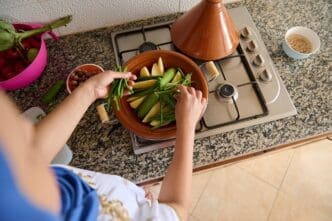 Top-down view of a woman adding fresh green beans to a steaming tagine pot filled with vegetables.