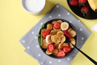 Overhead view of a dark bowl filled with tiny pancakes topped with strawberries and bananas, next to a cup of milk on a yellow background.