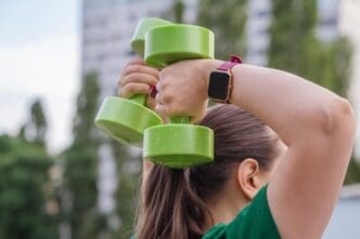 A woman in a green t-shirt does a dumbbell shoulder press exercise outdoors.