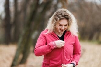 A woman in workout clothes prepares for a workout in a park during autumn, with colorful fall foliage visible.