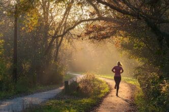 Woman running on a trail through a forest, with the sun rising in the background.