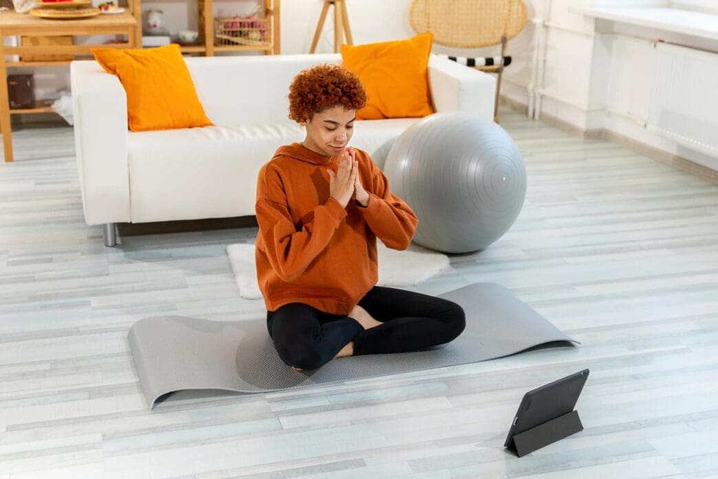 A young African girl sits in the lotus position on a yoga mat with her eyes closed and hands in prayer, meditating indoors.