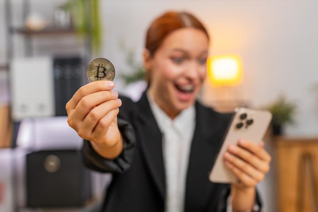Young businesswoman celebrates a successful investment by cheering at a smartphone displaying a rising Bitcoin chart.