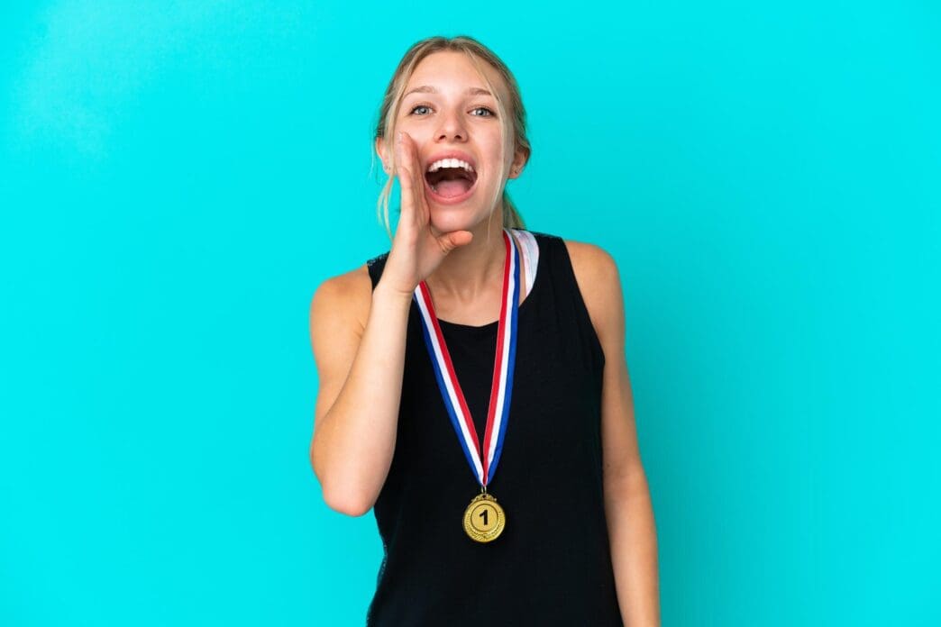 Young woman with fair skin wearing medals, isolated against a blue backdrop, shouts with her mouth open.