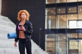 Young, red-haired European woman in athletic wear holds a phone and exercise mat outdoors.