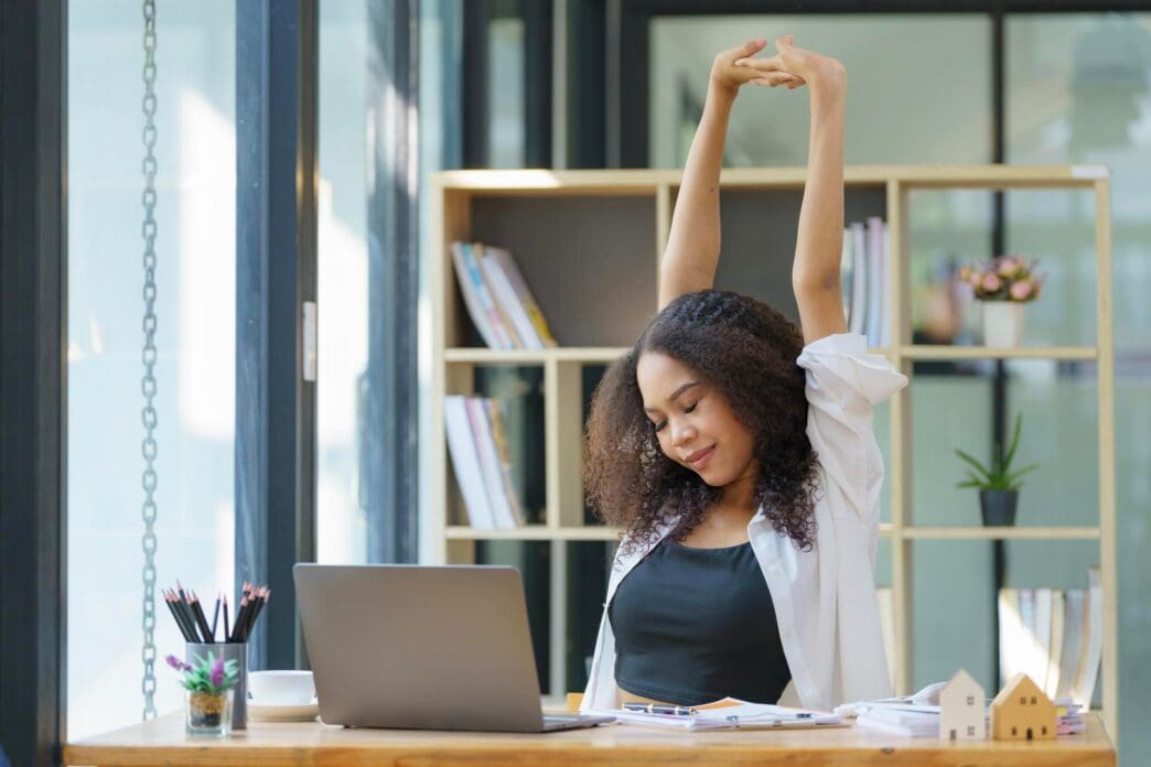 Young businesswoman stretches while working on a laptop at a modern workstation.