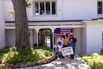 A multi-generational mixed-race family embraces a visitor in their garden, with children holding welcome signs and mini flags, and a large flag waving in the background.