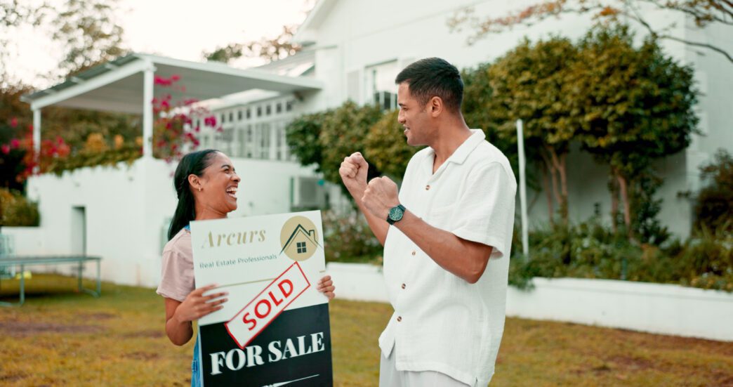 A smiling couple stands in front of their new home, holding a "sold" sign and celebrating their real estate investment.
