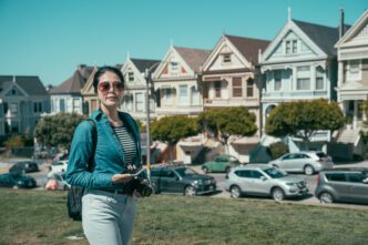 A young girl with a backpack and sunglasses uses her phone and camera while walking on a lawn in front of the colorful Painted Ladies Victorian houses in San Francisco.