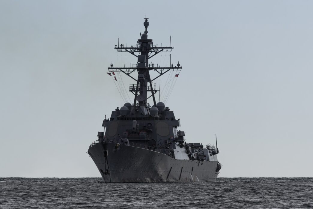 Front view of a large, gray American destroyer warship sailing on the dark, choppy sea under a pale sky.