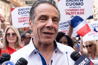 Former Governor Andrew Cuomo, wearing a white shirt, speaks into several microphones at a vibrant public parade.