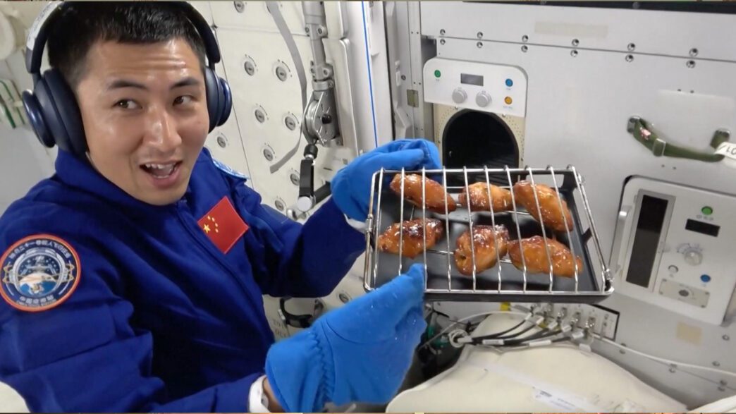 Chinese astronaut in space station holding a tray of baked chicken wings with a cheerful expression.