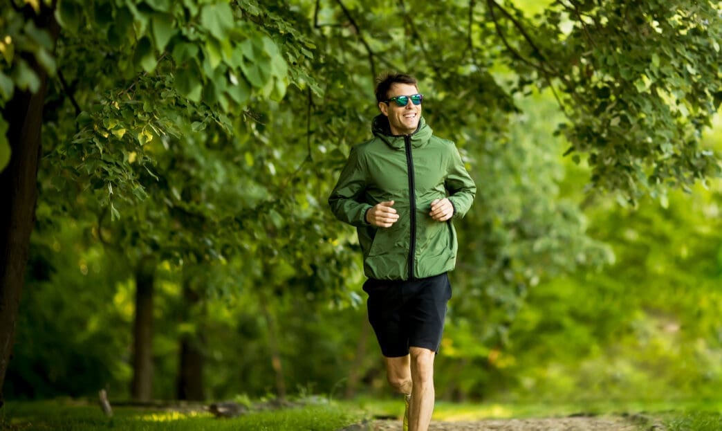 Young, athletic man in sunglasses and a green jacket running outdoors on a sunlit park path.