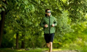Young, athletic man in sunglasses and a green jacket running outdoors on a sunlit park path.