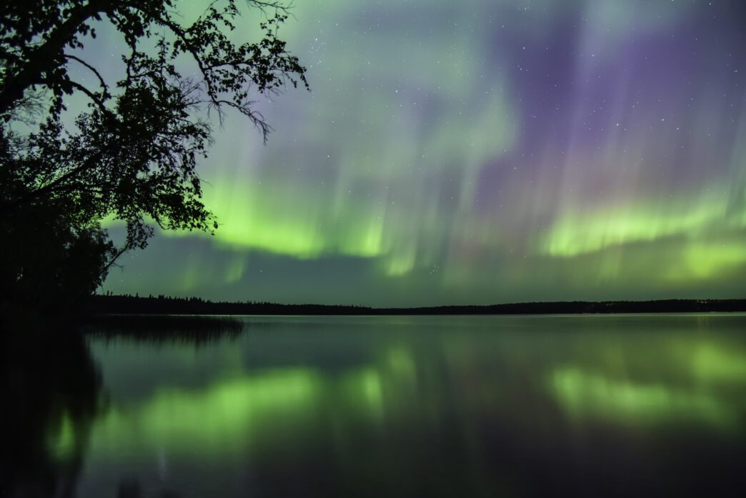 Vibrant green and purple Northern Lights reflecting dramatically on the smooth surface of a lake at night.