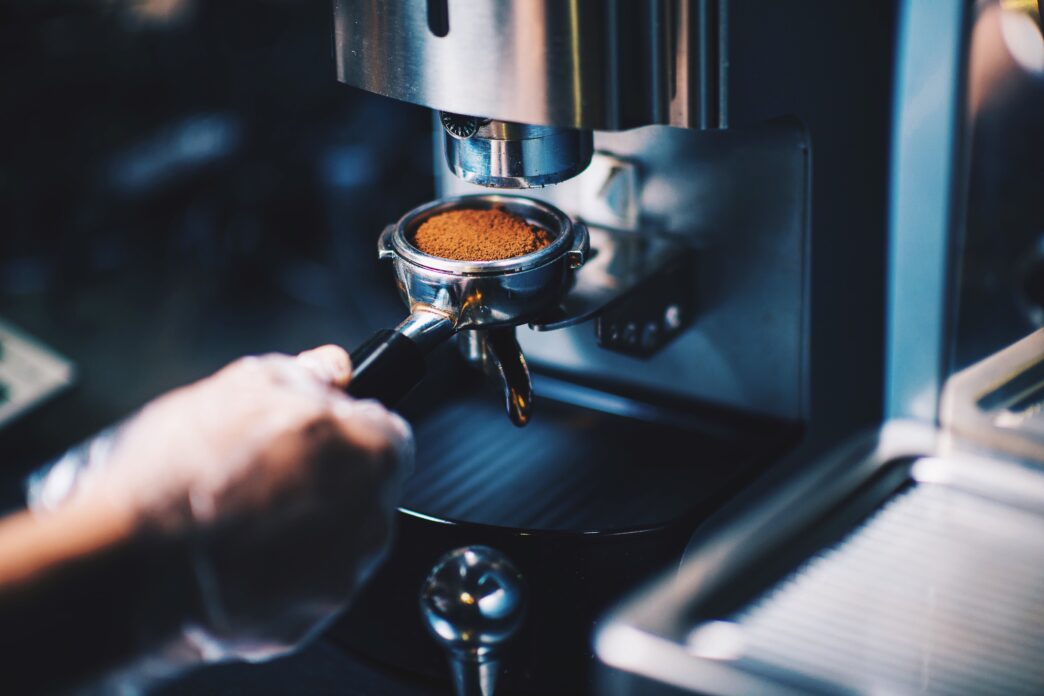 A barista's hand holding a portafilter filled with finely ground coffee under a coffee machine grinder.