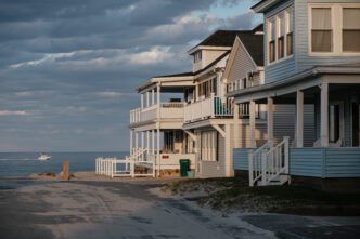 Three light blue and white wooden beach houses with multi-level porches stand on a sandy road at dusk.
