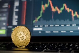 A golden physical Bitcoin coin resting on a laptop keyboard in front of a screen displaying a red and green trading chart.